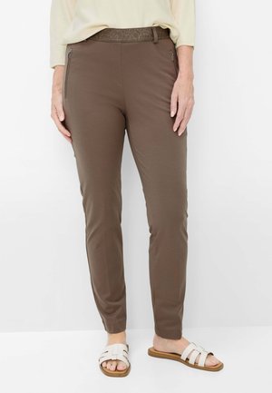 Kvinde iført brune tætsiddende bukser med lynlås-sidelommer, lysebluse og hvide åbne sandaler, stående mod en hvid baggrund.