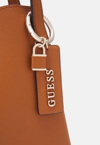 Sac à main en cuir marron avec une serrure dorée et une étiquette en cuir embossée du nom de la marque GUESS, attachée à une poignée anneau dorée.