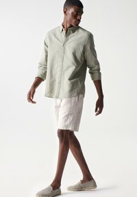 Camisa de botones verde claro, pantalones cortos de lino beige y zapatos gris claro sin cordones con suela trenzada. Modelo posando de manera casual contra un fondo blanco.