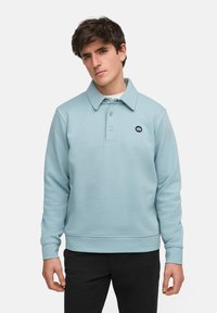 Ljusblå tröja med krage, tre knappar och en liten marinblå logotyp på bröstet. Har ribbade ärmmuddar och en texturerad tyg.