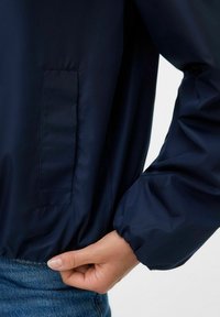 Asmuo reguliuoja elastingą juosmenį tamsiai mėlynos striukės, rodydamas rankovės apdailą ir stačiakampę siūtos kišenę šone.