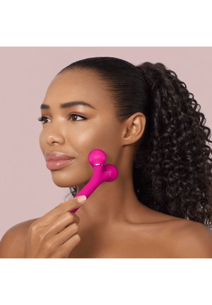 Femme aux cheveux bouclés utilisant un rouleau de massage facial rose sur sa joue sur un fond rose doux.