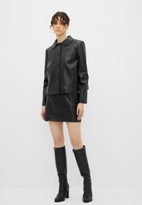 Schwarze Kunstlederjacke mit rundem Kragen und Knöpfen, kombiniert mit einem passenden Minirock und kniehohen Stiefeln mit Absatz. Glatte Textur.