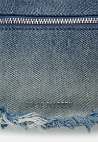 Denim torba s srebrnim zadrgo, strganim modrim robom in embosiranim logotipom "STEVE MADDEN". Ima teksturirano tkanino svetlo do srednje modre barve.