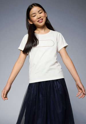 Witte t-shirt met opgerolde mouwen, voorzien van een textuur logo ontwerp. Gepaarde met een gelaagde, doorschijnende, donkerblauwe tule rok voor contrast.
