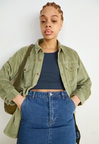 Grøn fløjlsskjorte, navy crop top og højtaljede blå denimnederdel med frontlommer. Glatte teksturer og stylet til et lag-på-lag-look.