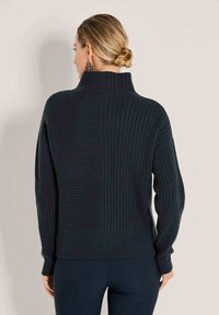 Frau mit blonden Haaren im Dutt, die einen schwarzen gerippten Pullover mit hohem Kragen trägt, von hinten betrachtet vor einem schlichten Hintergrund.