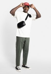 Black crossbody bag, white t-shirt, olive green pants, red cap with logo, black sneakers with white soles. Neutral background.