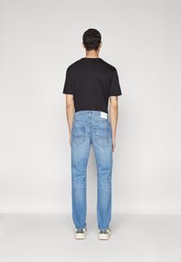 Îmbrăcăminte masculină care prezintă un tricou negru cu mânecă scurtă, blugi din denim albastru deschis și teniși bej cu accente verzi, văzuți din spate.