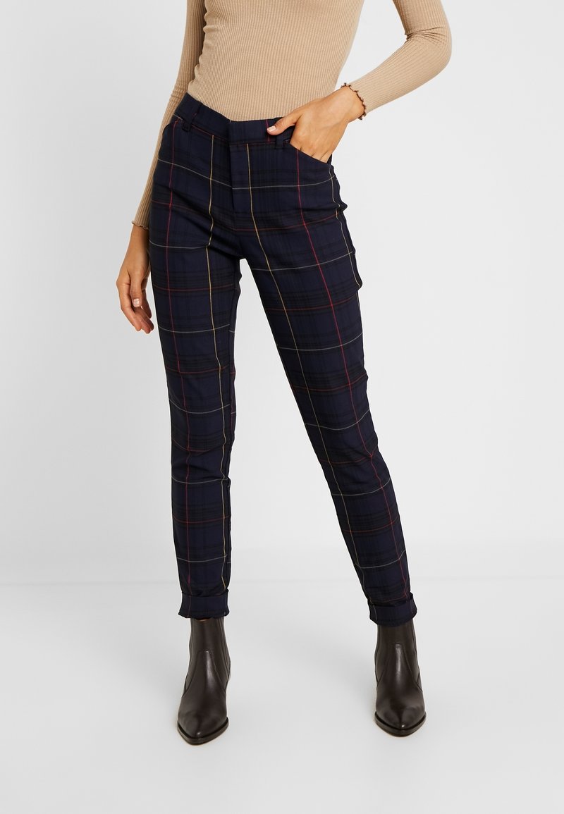 Femme portant un pantalon slim bleu marine à carreaux, un haut beige à manches longues en maille côtelée et des bottines marron foncé, les mains dans les poches.