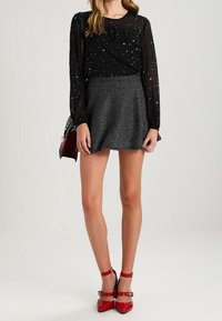 Haut noir transparent à manches longues orné d'étoiles argentées, associé à une jupe évasée grise. Talons aiguilles rouges vifs à lanières complètent la tenue.