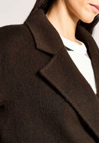 Eine Frau trägt einen dunkelbraunen Wollmantel mit breiten Revers über einem weißen Strickpullover, zu sehen von Hals bis Schulter.