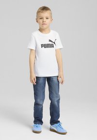 Balta T-kreka ar īsām piedurknēm, uz kuras ir melns 'PUMA' logo, kombinēta ar zilu džinsu biksēm un gaiši ziliem sporta apaviem. Vienkāršs dizains, kokvilnas materiāls.