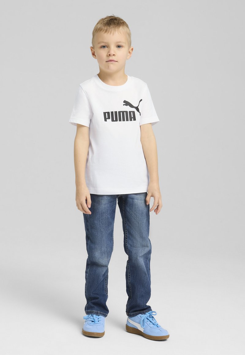 Balta T-kreka ar īsām piedurknēm, uz kuras ir melns 'PUMA' logo, kombinēta ar zilu džinsu biksēm un gaiši ziliem sporta apaviem. Vienkāršs dizains, kokvilnas materiāls.