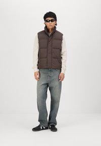 Brun polstret vest over en lys sweater, parret med lyseblå jeans og sorte sneakers. Vesten har vandret syninger og en høj krave.