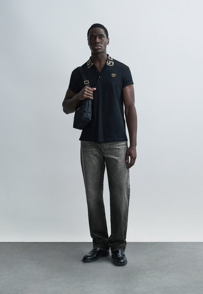 Juodas polo marškinėlis su auksu akcentuotu apykakle, pilkos džinsinės kelnės, juodi batai ir nedidelė siuvinėta peties rankinė. Minimalistinis fonas.