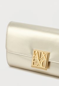 Μεταλλική χρυσή τσάντα αξεσουάρ με λεία υφή, διαθέτοντας κούμπωμα με τετράγωνο χρυσό λογότυπο. Ο σχεδιασμός είναι ορθογώνιος με καθαρές γραμμές και ραμμένα άκρα.