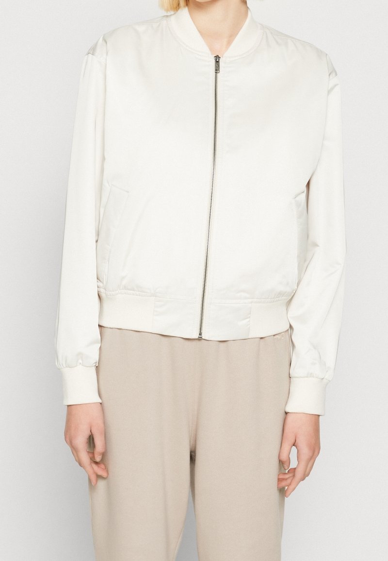 Světlý krémový bomber s hladkým materiálem, vybavený zipem vpředu, elastickými manžetami a bočními kapsami. Jednoduchý design bez vzorů.