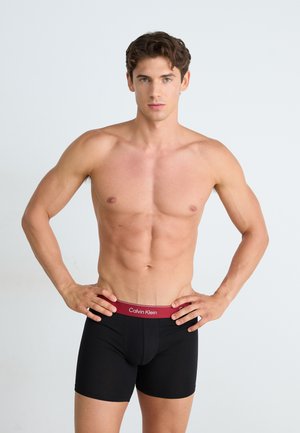 Zwarte boxershorts met een rode tailleband waarop het "Calvin Klein" logo staat. Gladde stof, aansluitend ontwerp en lengtemidden boven de dijen. Geen verdere details.