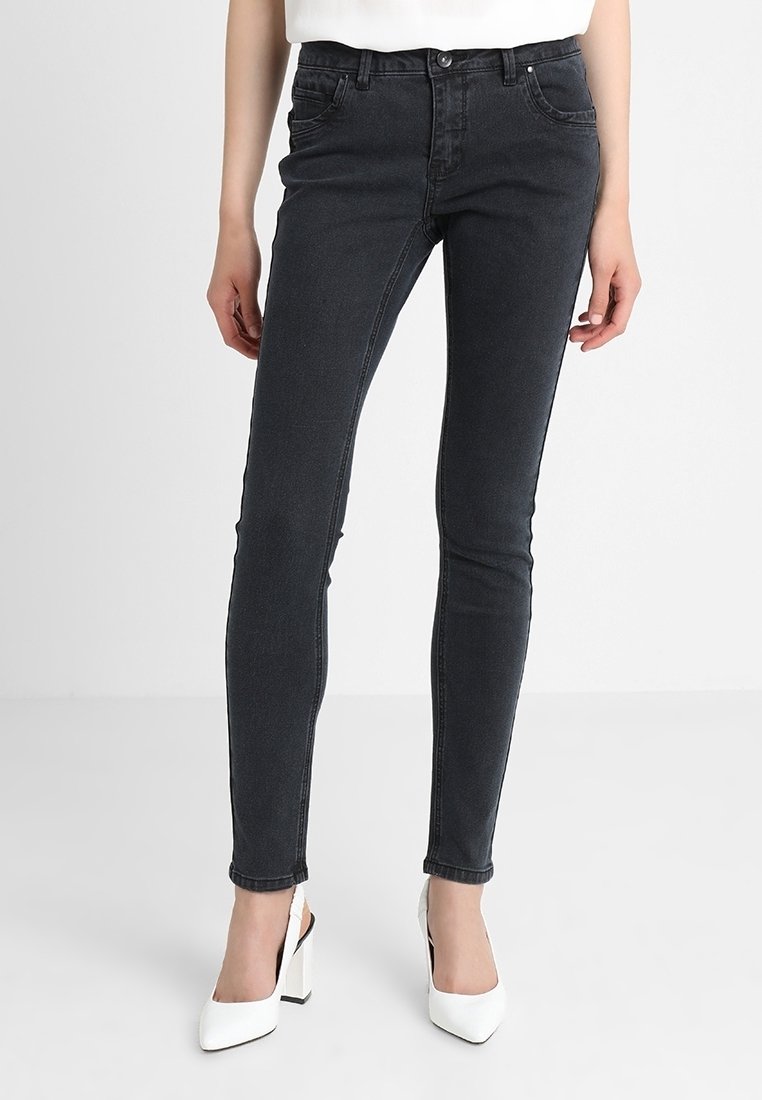 Dark gray skinny jeans made of denim, featuring a mid-rise waist, five-pocket design, and subtle stitching details. Worn with white heels.