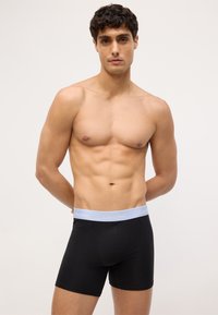 Čierne bavlnené boxerky s elastickým pásom svetlomodrej farby, na ktorom je logo "Calvin Klein", zobrazené na modeli s neutrálnym pozadím.