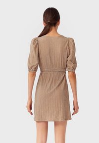 Femme aux cheveux bruns en queue de cheval basse, portant une robe beige à pois avec des manches bouffantes, se tenant de dos sur un fond blanc.