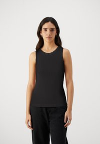 Svart ermeløs ribbestrikket topp med rund hals, med en tettsittende design som strekker seg til hoftene. Kombinert med mørke, løse bukser.