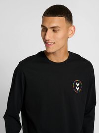 Młody mężczyzna z krótkimi włosami, noszący czarną koszulkę z długim rękawem z małym okrągłym logo i napisem na lewej piersi.