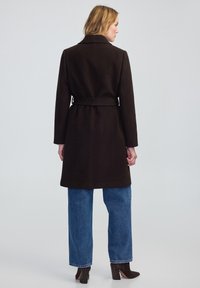 Cappotto in misto lana marrone con vita aderente, rever a punta e maniche lunghe, indossato sopra jeans a gamba larga blu e stivaletti con tacco.