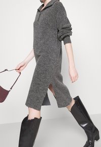 Robe tricotée grise avec un col zippé, fentes latérales et manches longues. Portée avec des bottes noires jusqu'aux genoux et tenant un sac à main bordeaux.