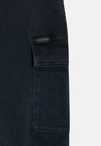Pantalones cargo de denim oscuro con un gran bolsillo lateral que presenta una etiqueta tejida de Calvin Klein. La tela tiene una textura suave y un tono azul profundo.