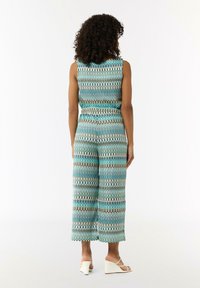 Jednoczęściowy kombinezon bez rękawów w kolorze teal, z falistym wzorem w odcieniach turkusu, kremu i brązu. Luźny krój z szerokimi nogawkami i elastycznym paskiem w talii. Noszony z białymi sandałami na koturnie.