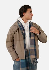 Giacca marrone chiaro con zipper e una texture liscia, indossata sopra un hoodie bianco. Sciarpa a quadri blu con bordi frangiati appoggiata intorno al collo.