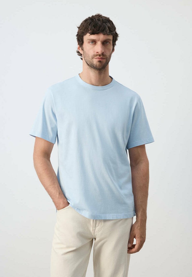 Homme aux cheveux courts et foncés, portant un t-shirt bleu clair et un pantalon beige, debout devant un fond clair uni, une main dans la poche.