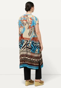 Tunika a maniche corte, ampia, di colore beige con motivi astratti blu e marroni, caratterizzata da spacchi laterali e orlo dritto. Indossata sopra pantaloni neri.