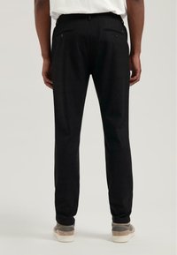 Schwarze Hose mit geradem Bein, ausgestattet mit zwei Gesäßtaschen und einer glatten Textur. Kombiniert mit grau-weißen Schuhen.