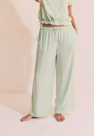 Vrouw die lichtgroene en witte geruite broek met wijde pijpen en bijpassende cropped top draagt, blootsvoets staand op een lichte vloer.