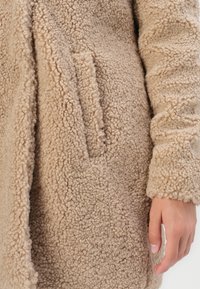 Béžový kabát z umělé kožešiny s měkkým, texturovaným povrchem, který má skrytou kapsu a dlouhé rukávy. Teplý a útulný design ideální pro chladnější počasí.