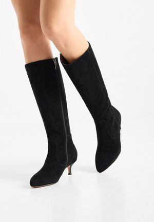 Gambe che indossano stivali al ginocchio in camoscio nero con zip laterale e tacchi bassi kitten, posate su sfondo bianco.