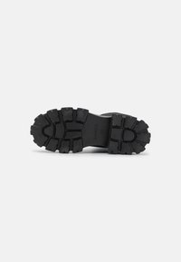Suela de bota de goma negra con un patrón de relieve profundo, que presenta una superficie texturizada y un nombre de marca en relieve. Tamaño marcado como 37.