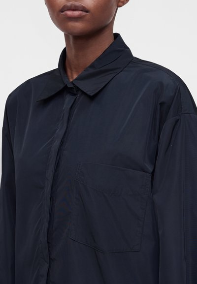 Personne portant une chemise bleu marine foncé à col avec une grande poche poitrine, montrée des épaules juste au-dessus des lèvres.