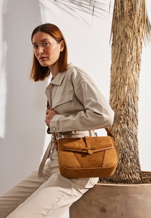 Mujer con cabello rojo liso, vestida con chaqueta y pantalones beige, sentada junto a una palmera en maceta, sosteniendo un bolso de ante color beige.