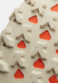 Gros plan sur la semelle beige d'une chaussure avec des crampons en forme de flèches tridimensionnelles et des découpes triangulaires orange pour l'adhérence.