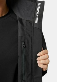 Chaqueta negra con una textura suave y forro de polar, que cuenta con un bolsillo lateral con cremallera y el logo "Helly Hansen" en el borde del cuello.
