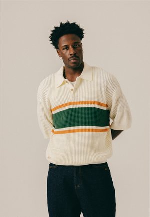 Homme aux cheveux courts et bouclés portant un pull en maille crème avec des rayures vertes et orange, debout avec les mains derrière le dos devant un fond uni.