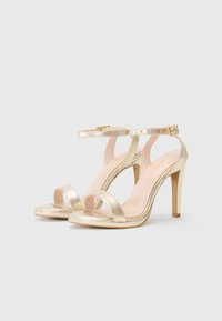 Goldfarbene, metallische High Heels mit zwei Riemen über den Zehen, einem Knöchelriemen mit Schnalle und einem schmalen, sich verjüngenden Absatz.