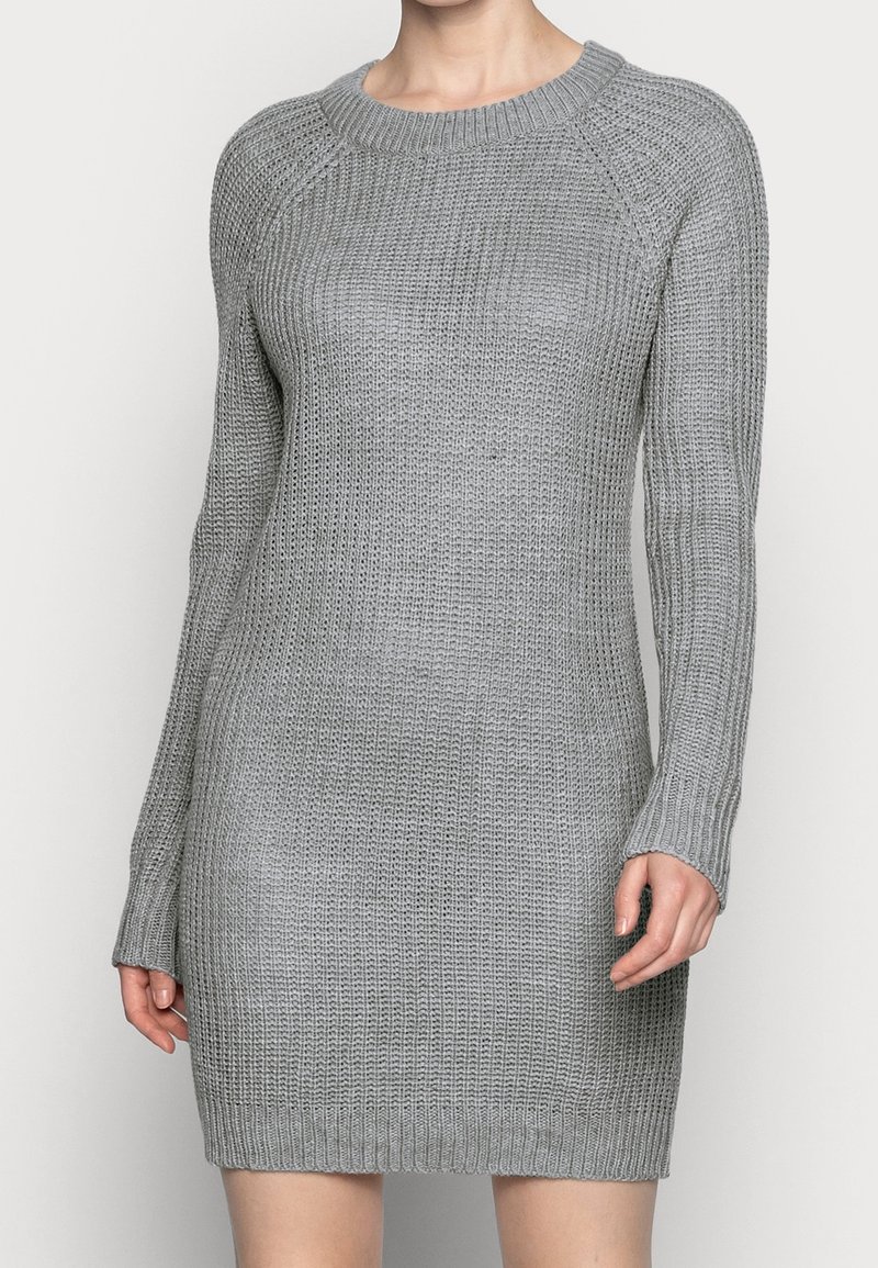 Femme portant une robe pull grise claire, tricot épais à manches longues et col rond, debout devant un arrière-plan uni.