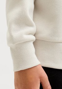 Svetlo sivi spodnji rob jopice z rebrasto teksturo. Mehka tkanina s subtilnimi madeži, ki poudarja oprijet dizajn okoli zapestja.