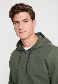 Groene rits hoodie met een trekkoordkap, zwart-witte patroonkoord en een gladde textuur. Draagt over een wit shirt.
