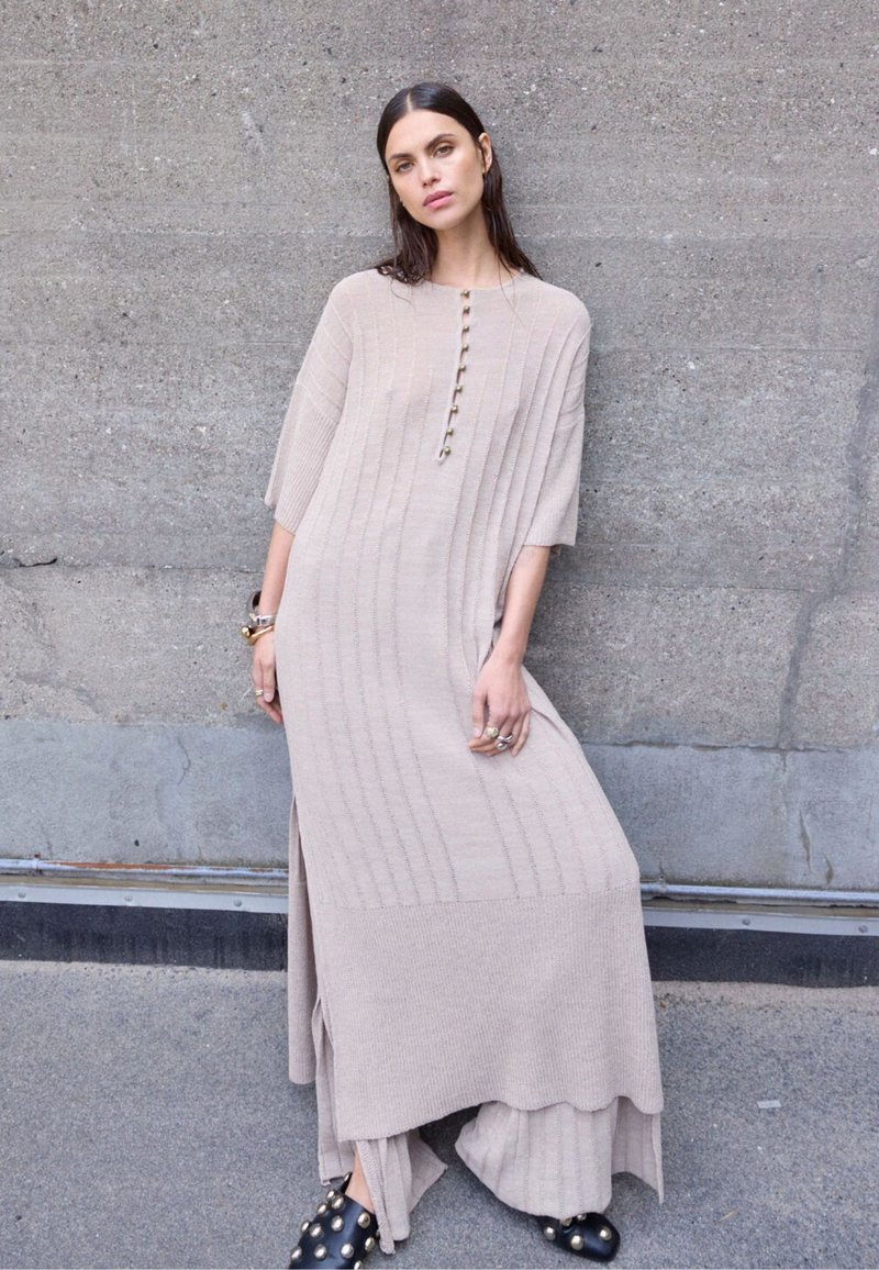 Μπεζ πλεκτό φόρεμα με κοντά μανίκια, κάθετη ραβδώσεις, λεπτομέρειες με κουμπιά, πλαϊνές σχισμές, σχεδιασμός με στρώσεις πάνω σε φαρδιά παντελόνια και μαύρα χοντρά παπούτσια με καρφιά.